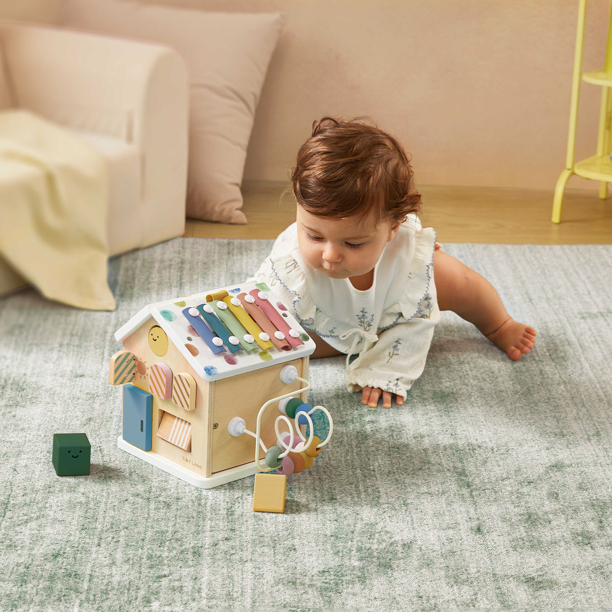A baby in white lies on a soft gray Cozy Learning Playset — Playmat by Tiny Land, playing with the included activity cube featuring a xylophone and shapes. A green block from the set is nearby.