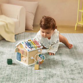 A baby in a white outfit plays on a soft rug with the Tiny Land® Cottage Activity Cube - Morandi by Tiny Land, a wooden house-shaped Montessori toy featuring a xylophone roof and sensory elements for developmental play.
