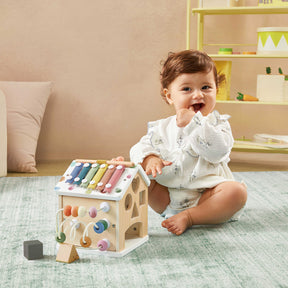 A smiling baby in a white outfit plays on the carpet with the Tiny Land® Cottage Activity Cube - Morandi by Tiny Land, shaped like a house, while shelves and toys are visible in the background.