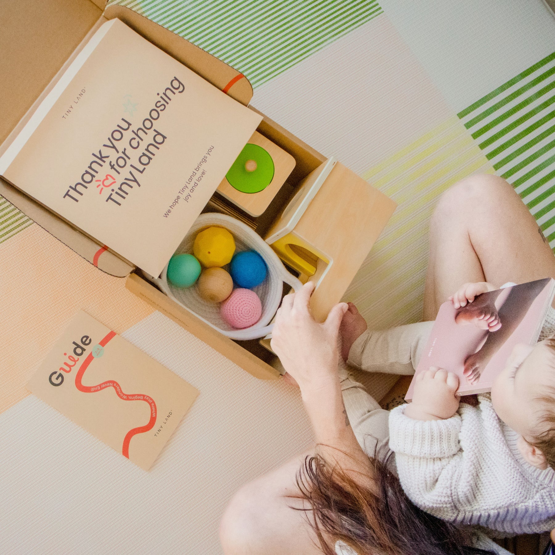 A woman and her baby sit on a pastel play mat, opening the Tiny Land® Montessori Early Skills Baby Set (7–12 months). A card says “Thank you for choosing Tiny Land,” with a guide booklet nearby.
