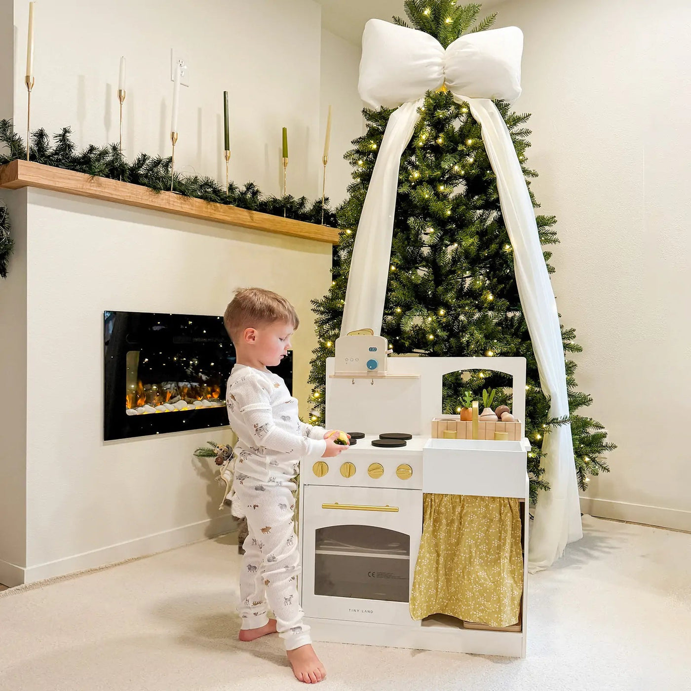 A young child in pajamas enjoys imaginative play with the Tiny Land® Wooden Play Kitchen for Toddlers – Honey Kitchenette Pretend Cooking Set, surrounded by Christmas decor including a lit tree with a white bow and a mantle adorned with greenery and candles.