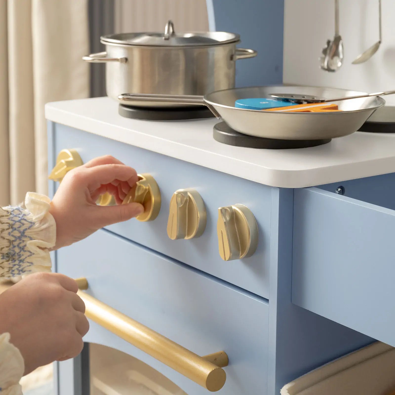 A child turns a gold knob on the blue Tiny Land® Serenity Wooden Kitchen & Wooden Food Set, with pots and pans on the burners, making this Tiny Land toy a charming playroom centerpiece.
