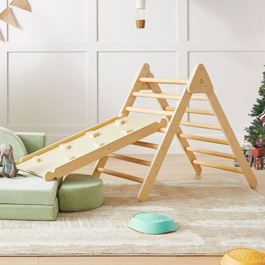 The Tiny Land® Pikler Triangle 7-in-1 Montessori Climbing Set sits on a rug in the living room near a plush elephant toy, a green sofa, and a decorated Christmas tree.|natural wood color