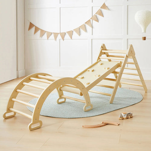 The Tiny Land® Pikler Triangle 7-in-1 Montessori Climbing Set by Tiny Land, featuring a triangle, arch ladder, and ramp with climbing holds, is displayed on a light blue rug in a bright room with wooden floors and playful decor.