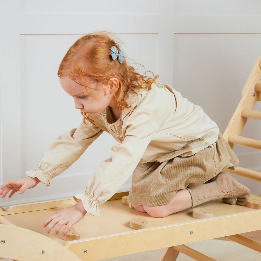 A young girl in beige and brown explores the Tiny Land® 7-in-1 Montessori Climbing Set with Play Pad indoors, kneeling on the wooden structure with a blue bow in her red hair, fully engaged with the Tiny Land play equipment.|natural wood color