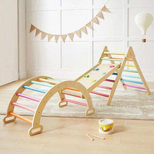 The Tiny Land® 7-in-1 Rainbow Climbing Set by Tiny Land includes a Pikler Triangle, wooden arch ladder, and slanted climbing board with colorful grips. A toy drum and stick also sit on the light wood floor in this bright room.