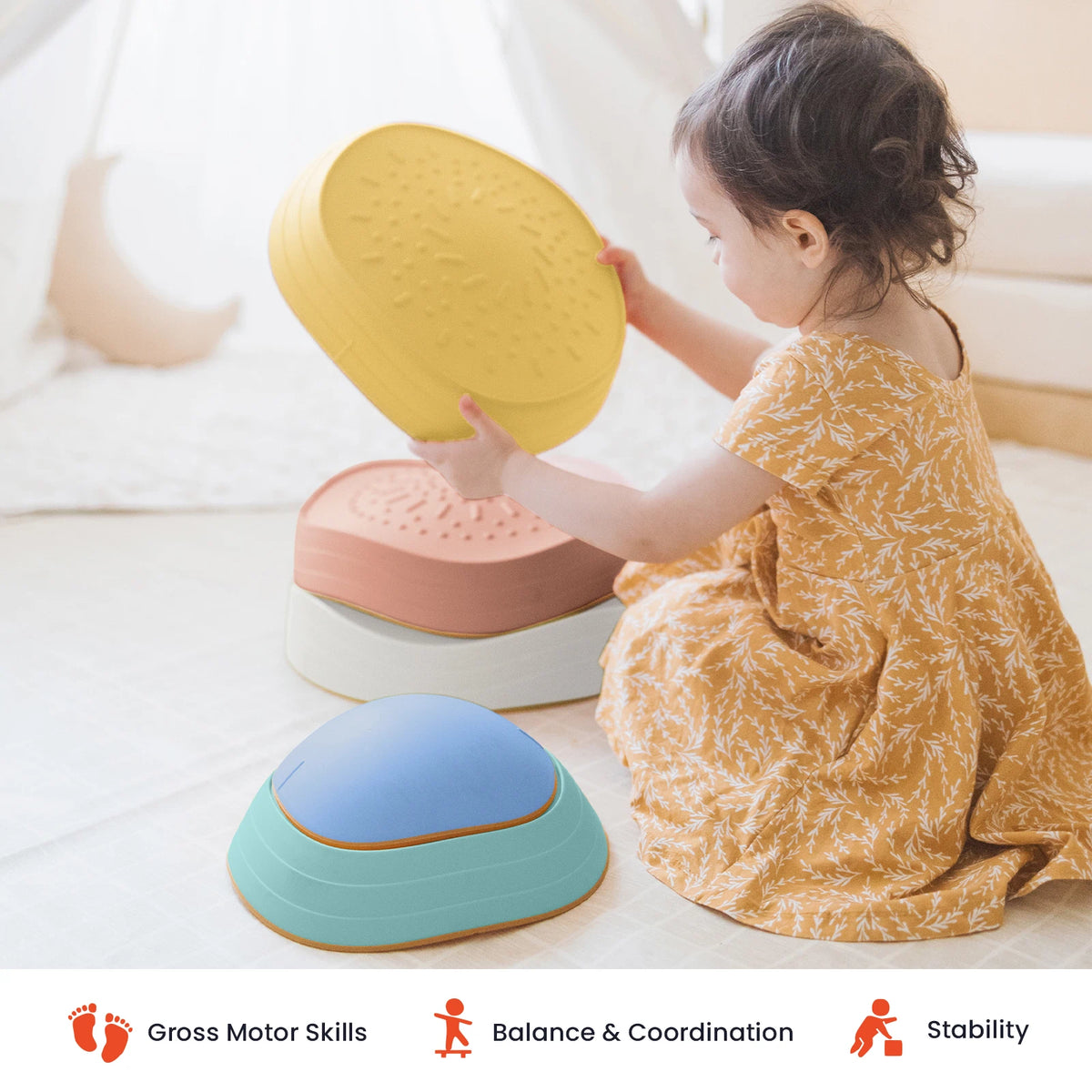 A young child in a yellow dress stacks Tiny Land® Stepping Stones - Beach Vacation by Tiny Land in a bright room. Icons and text below highlight benefits: motor skills development, balance & coordination, and stability—making these stepping stones perfect for creating a fun obstacle course at home.