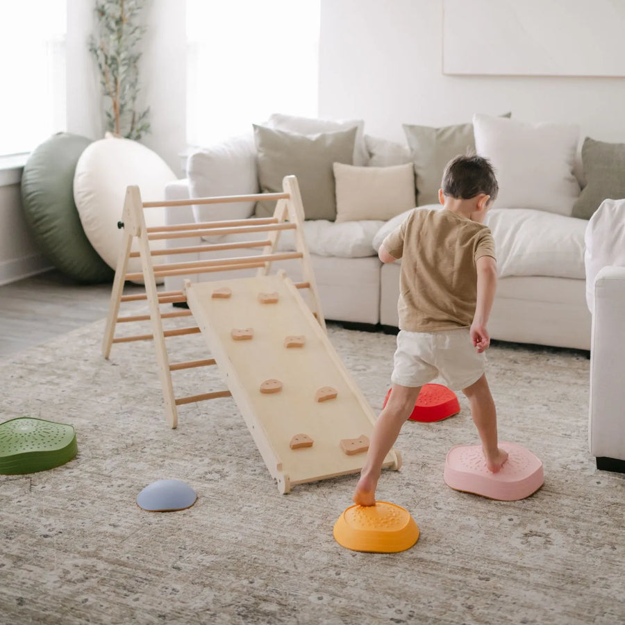 A young child stands barefoot on Tiny Land® Stepping Stones - Vintage Color in a bright living room, facing a wooden climbing ramp. With pillows and a couch nearby, the space creates an inviting Tiny Land motor skills obstacle course for indoor play.|Vintage Color