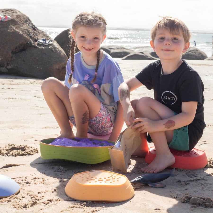 Two young children build motor skills at the beach using Tiny Land® Stepping Stones - Vintage Color. The girl in a purple shirt and the boy in black play as the ocean stretches behind them, enjoying their Tiny Land activity together.|Vintage Color