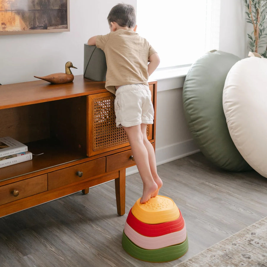 A young boy uses Tiny Land® Stepping Stones - Vintage Color from Tiny Land to reach a gray box atop a wooden cabinet, enhancing his motor skills in a bright, modern living room.|Vintage Color