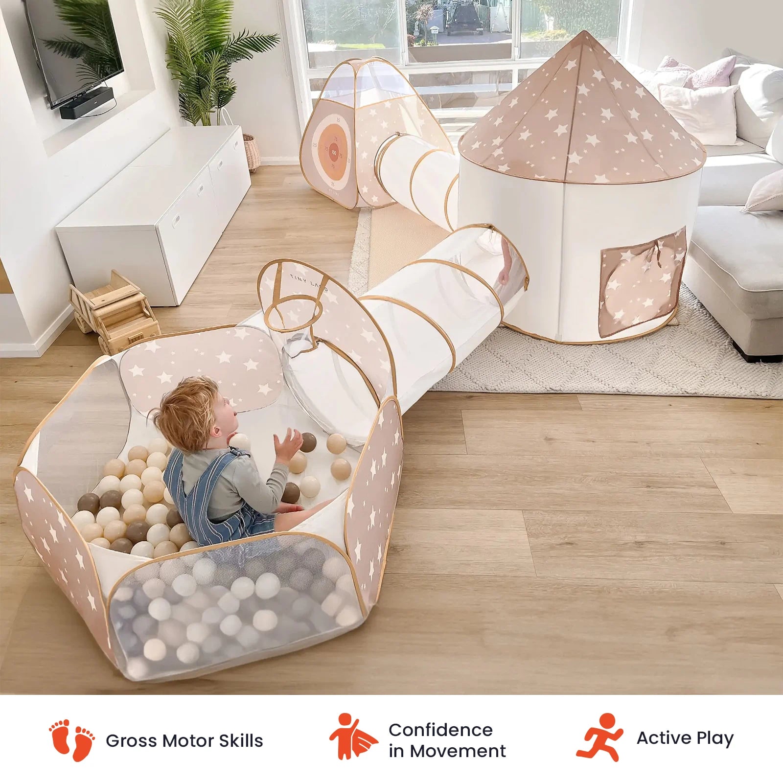 A young child plays in the Tiny Land® Discovery 5-in-1 Play Tunnel & Ocean Ball Set by Tiny Land—a beige and white ball pit complete with an adventure tunnel and play tent—inside a bright living room. Icons below highlight benefits: gross motor skills, confidence in movement, and active play.
