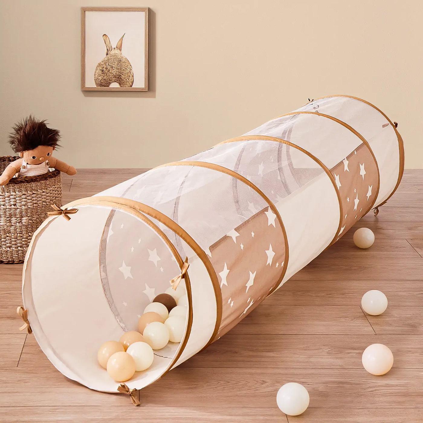 The Tiny Land® Adventure Play Tunnel, featuring mesh fabric and white stars, sits on a wooden floor with soft play balls. A plush doll in a woven basket and framed animal art decorate the beige wall in the background.