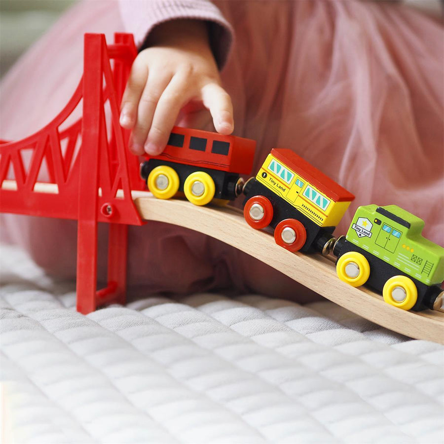 A child in a pink top and skirt pushes colorful trains over a red bridge from the Tiny Land® Wooden Train Set 110 Pcs by Tiny Land, compatible with all major brands, on a quilted surface.|110 Pcs