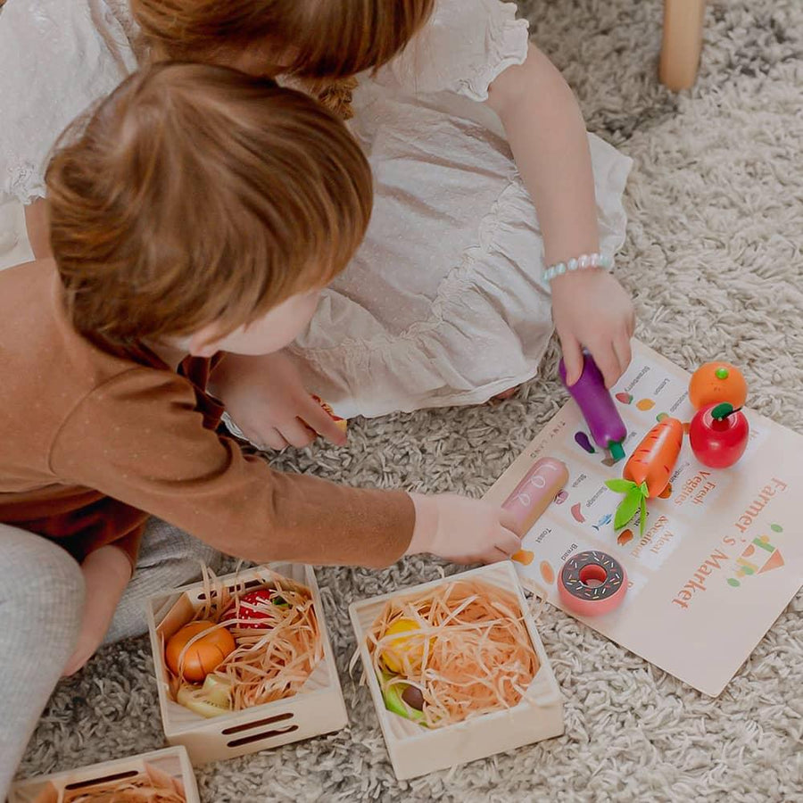 Two kids sit on a carpet, playing with the Tiny Land® Wooden Food for Play Kitchen & Wooden Rack by Tiny Land Play Food. They arrange realistic wooden play food in crates and point to a Farmers Market menu on the floor.