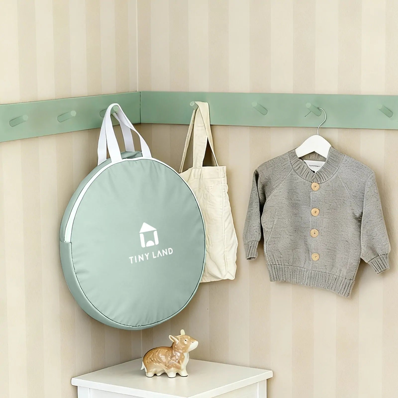 A soft green round bag labeled Tiny Land, a beige tote, and a gray knit sweater hang above a white shelf with a ceramic cow—charming decor for cozy indoor adventures with the Tiny Land® Tiny Sprout Castle Tent.
