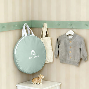 A soft green round bag labeled Tiny Land, a beige tote, and a gray knit sweater hang above a white shelf with a ceramic cow—charming decor for cozy indoor adventures with the Tiny Land® Tiny Sprout Castle Tent.