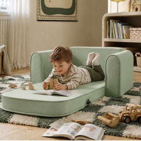 A young child relaxes on the Tiny Land® GentleSnug Kids Sofa Chair by Tiny Land, reading a book and playing with wooden blocks in a cozy, well-lit room filled with shelves, toys, and books.