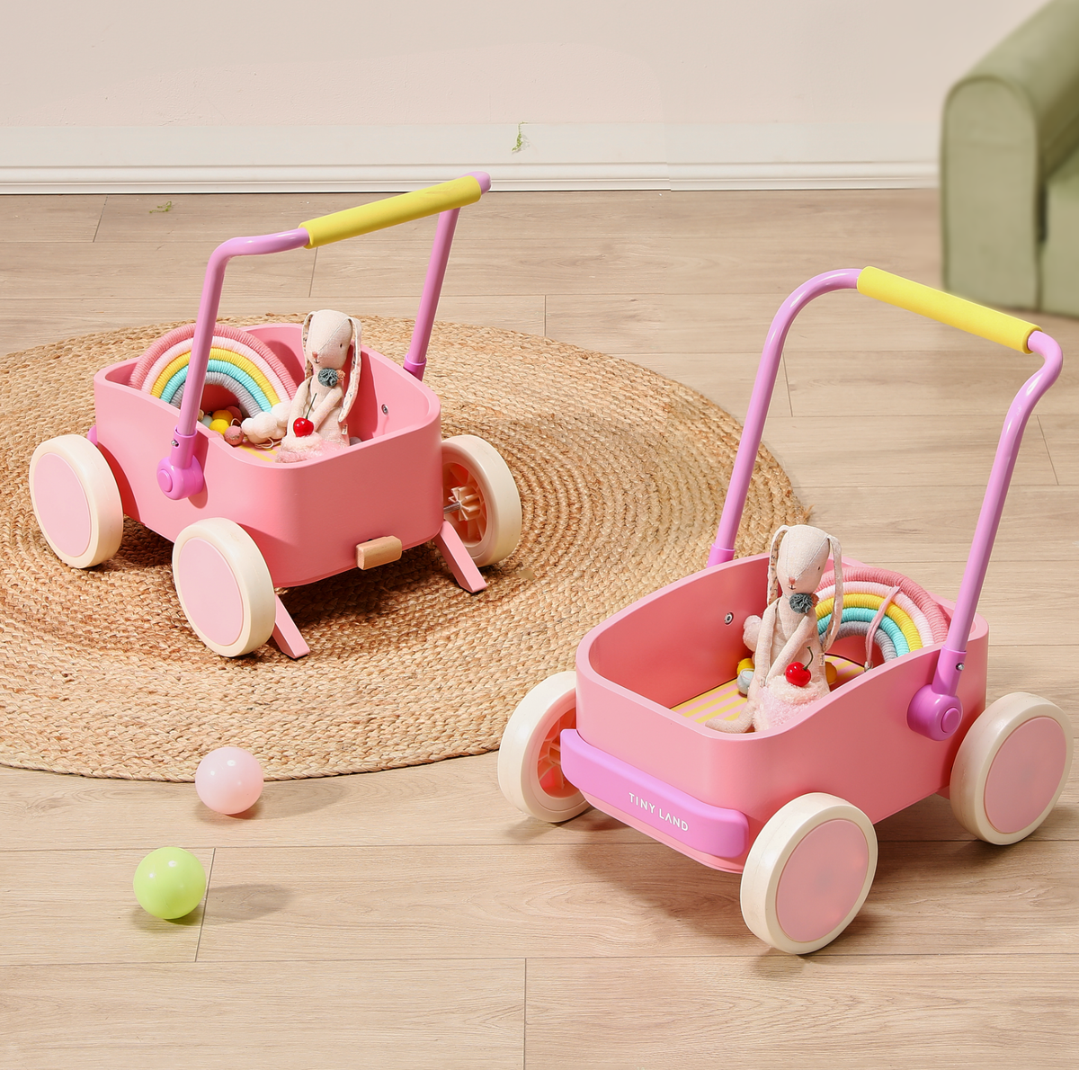Two Test Tiny Land® JoyRide Baby Walker with Blocks - Pink, each holding a stuffed animal and a rainbow toy, are on a woven round rug on a wooden floor. Colorful balls are scattered nearby, with part of a green couch visible.