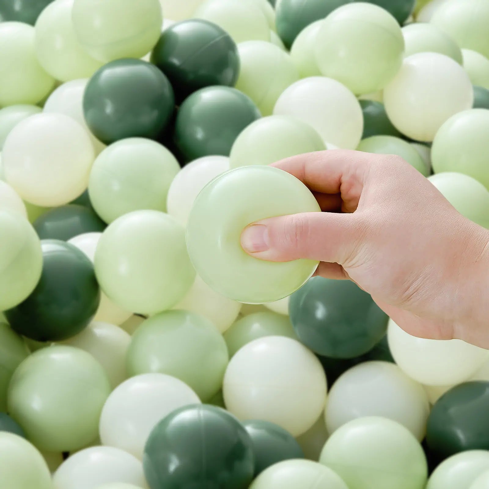 A hand holds a Tiny Land® Green Ocean Ball over a pile of safe, non-toxic play balls in light green, dark green, and off-white from the 200-piece Tiny Land® Green Ocean Ball set.