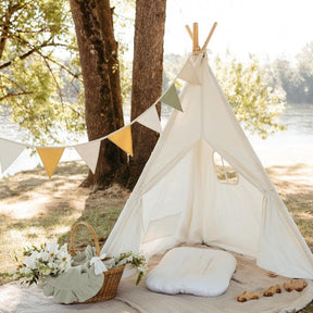 The Tiny Land® Teepee Tent for Kids with Mat by Tiny Land is set up outdoors on a blanket near trees and a lake. The white fabric play tent, featuring a cozy floor mat inside, is decorated with a basket of white flowers and a string of yellow and green pennant flags.