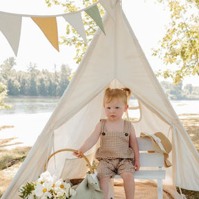 A toddler with pigtails sits on a white bench inside the Tiny Land® Teepee Tent for Kids with Mat, a sturdy play tent by Tiny Land designed in a charming small teepee shape. Set outdoors, she wears a checked romper and is surrounded by flowers, a sun hat, and a stuffed toy. The picturesque scene includes trees and a river in the background.