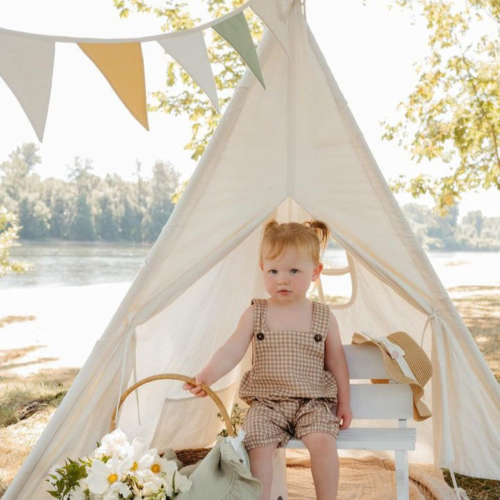 A toddler with pigtails sits on a white bench inside the Tiny Land® Teepee Tent for Kids with Mat, a sturdy play tent by Tiny Land designed in a charming small teepee shape. Set outdoors, she wears a checked romper and is surrounded by flowers, a sun hat, and a stuffed toy. The picturesque scene includes trees and a river in the background.