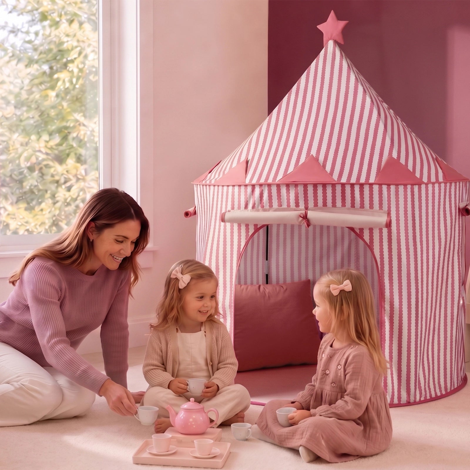 A woman and two young girls enjoy a tea party on a pink carpet next to the Tiny Land® Kids Tent – an indoor tent for toddlers by Tiny Land, which features playful pink stripes. All three are dressed in pastel clothes and smile happily as sunlight pours through a large window.|PInk