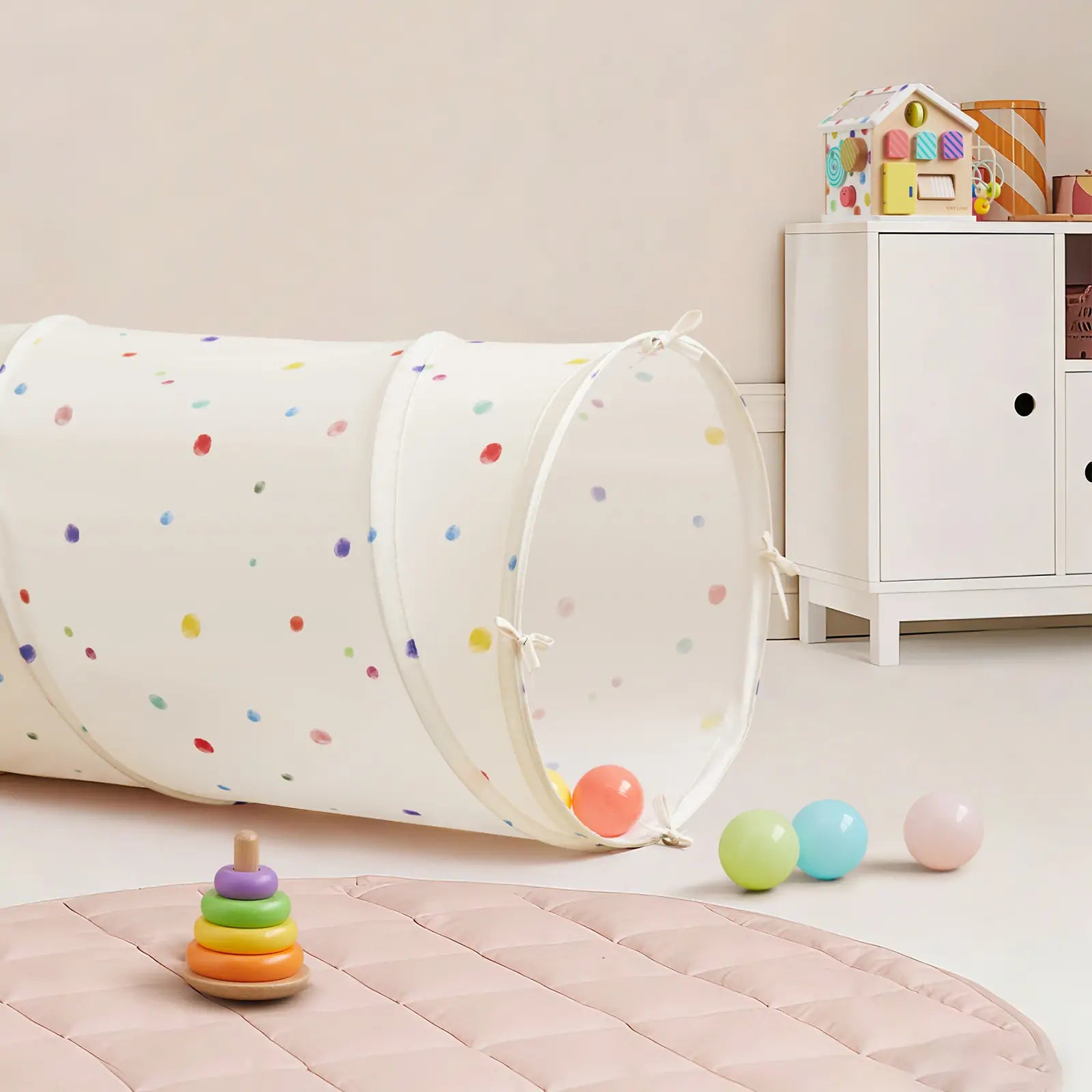 A children's playroom featuring the Tiny Land® Adventure Play Tunnel—Colorful Polka Dots, pastel toy balls, a rainbow ring stacker, a padded pink play mat, a white cabinet, and a colorful toy house on top of the cabinet.