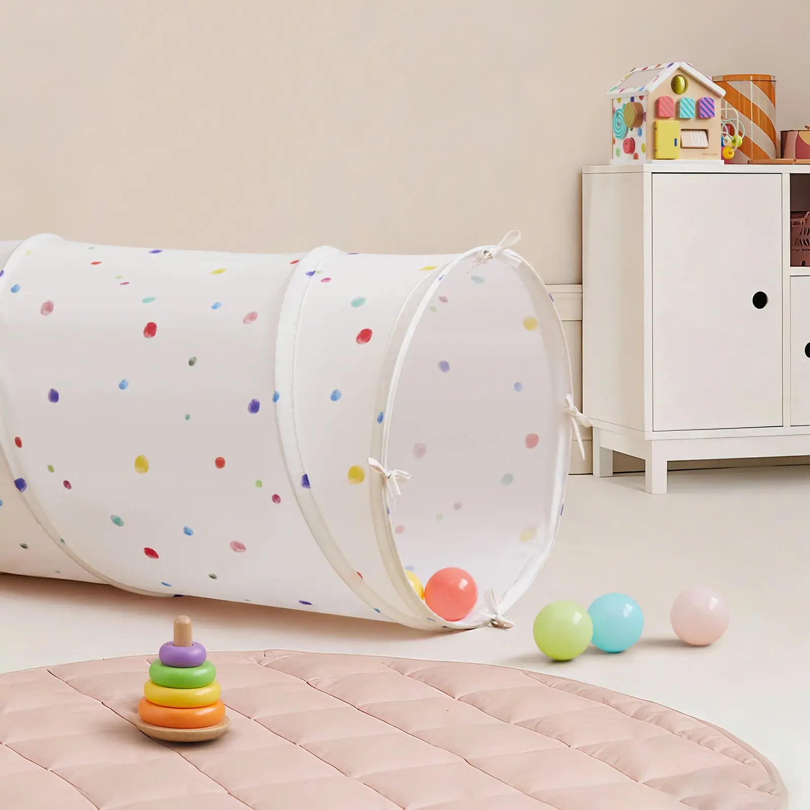 A cheerful children’s playroom featuring the Tiny Land® Adventure Play Tunnel - Colorful Polka Dots, scattered balls, a ring stacking toy on a pink mat, and a white cabinet with a toy house in the background.|white