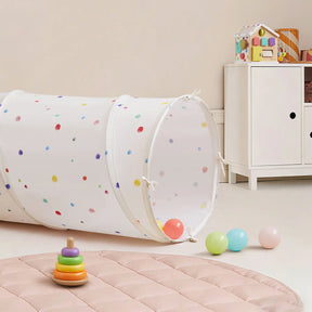 A cheerful children’s playroom featuring the Tiny Land® Adventure Play Tunnel - Colorful Polka Dots, scattered balls, a ring stacking toy on a pink mat, and a white cabinet with a toy house in the background.|white