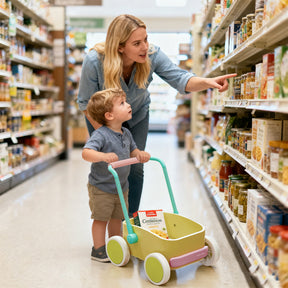 A woman points to a shelf in a grocery store aisle while a young child stands nearby pushing a pink and spring-colored Tiny Land® 3-in-1 Wooden Baby Walker Wagon with an integrated storage box.|Spring Color