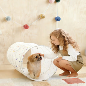 A young girl with curly hair crouches beside the Tiny Land® Adventure Play Tunnel - Colorful Polka Dots, gently petting a small brown and white puppy inside. Bright pom-poms hang on the wooden wall, adding to the playful scene.|white