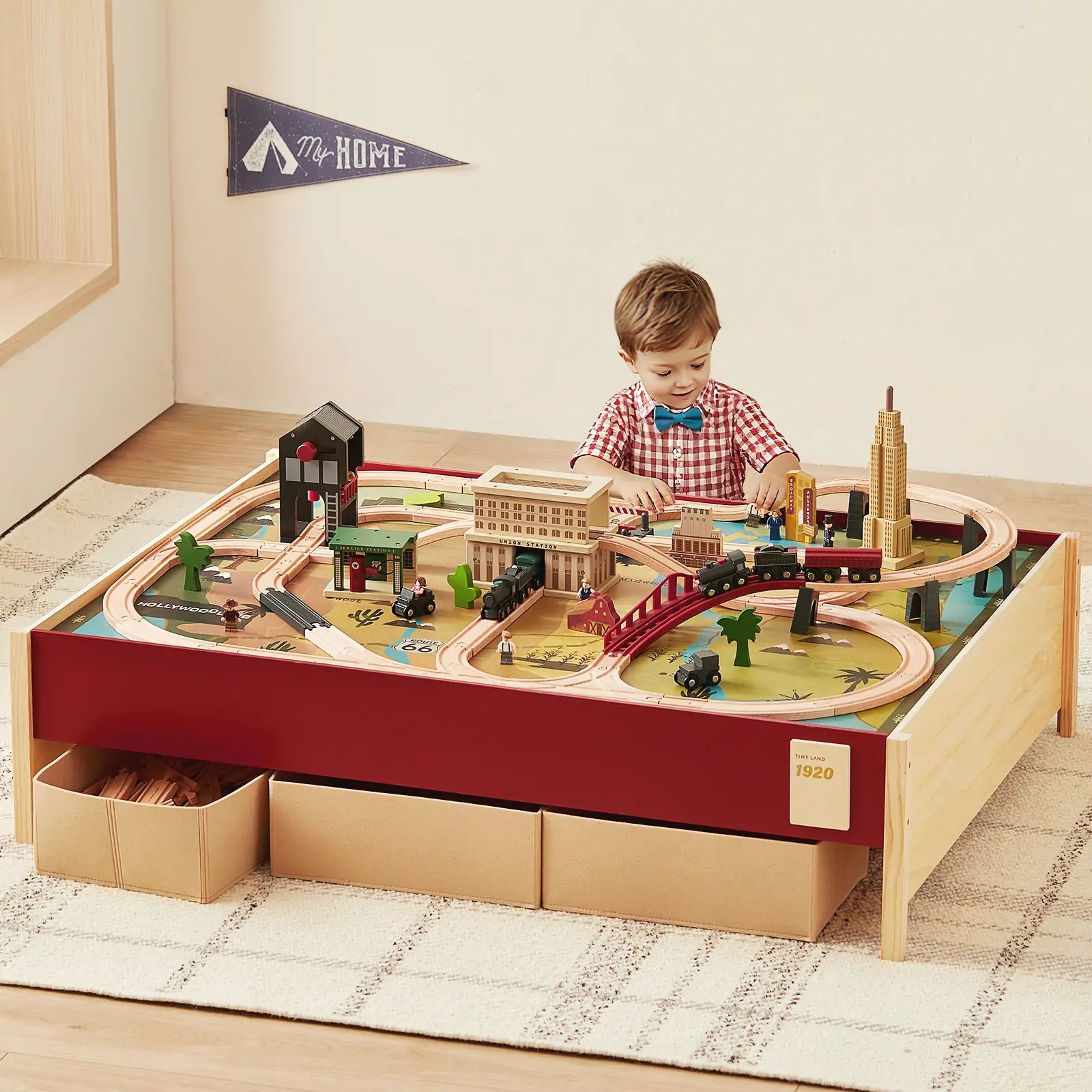 A young child plays with the Tiny Land® 1920 Railway Adventure Train Table by Tiny Land INC, surrounded by tracks, buildings, and vehicles. Storage boxes sit below the table, and a "My Home" pennant hangs on the wall behind.