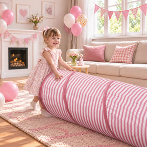 A smiling young girl in a pink dress plays in her decorated living room with the Tiny Land® Sweetwood Adventure Dollhouse & Play Tunnel Set, featuring a long, pink-and-white striped cylindrical play tunnel.