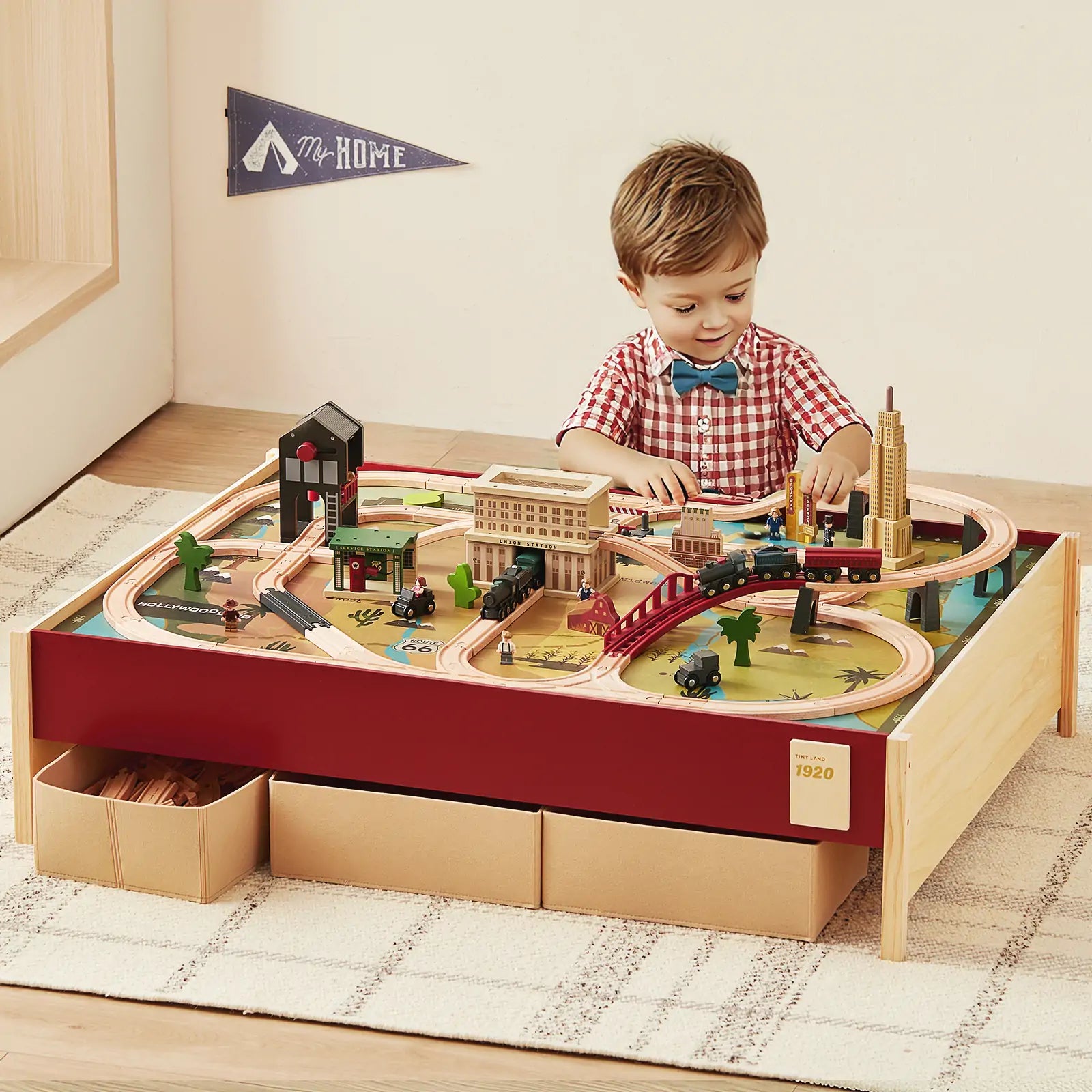 A young boy plays with the Tiny Land® 1920 Railway Adventure Train Table by Tiny Land INC, featuring built-in storage boxes. The table sits in a cozy room with a rug and a “My Home” wall decoration.