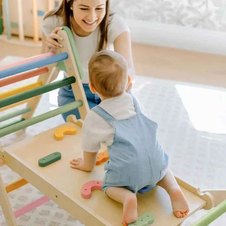 A baby in blue overalls climbs the Tiny Land® 7-in-1 Morandi Pikler Triangle Set by Tiny Land, using colorful handles as an encouraging adult smiles nearby in a bright, child-friendly room.