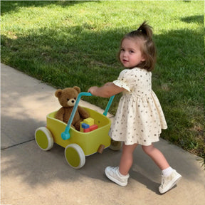 A young girl in a white dress pushes the Test Tiny Land® JoyRide Baby Walker with Blocks - Spring Color, carrying a teddy bear and blocks, along a sidewalk beside green grass.