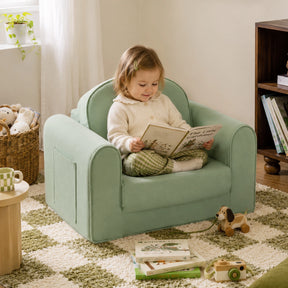 A young child sits cross-legged on the Tiny Land® GentleSnug Kids Sofa Chair, smiling as they read a book. The cozy room is filled with wooden toys, books, a soft rug, and natural light from a nearby window.