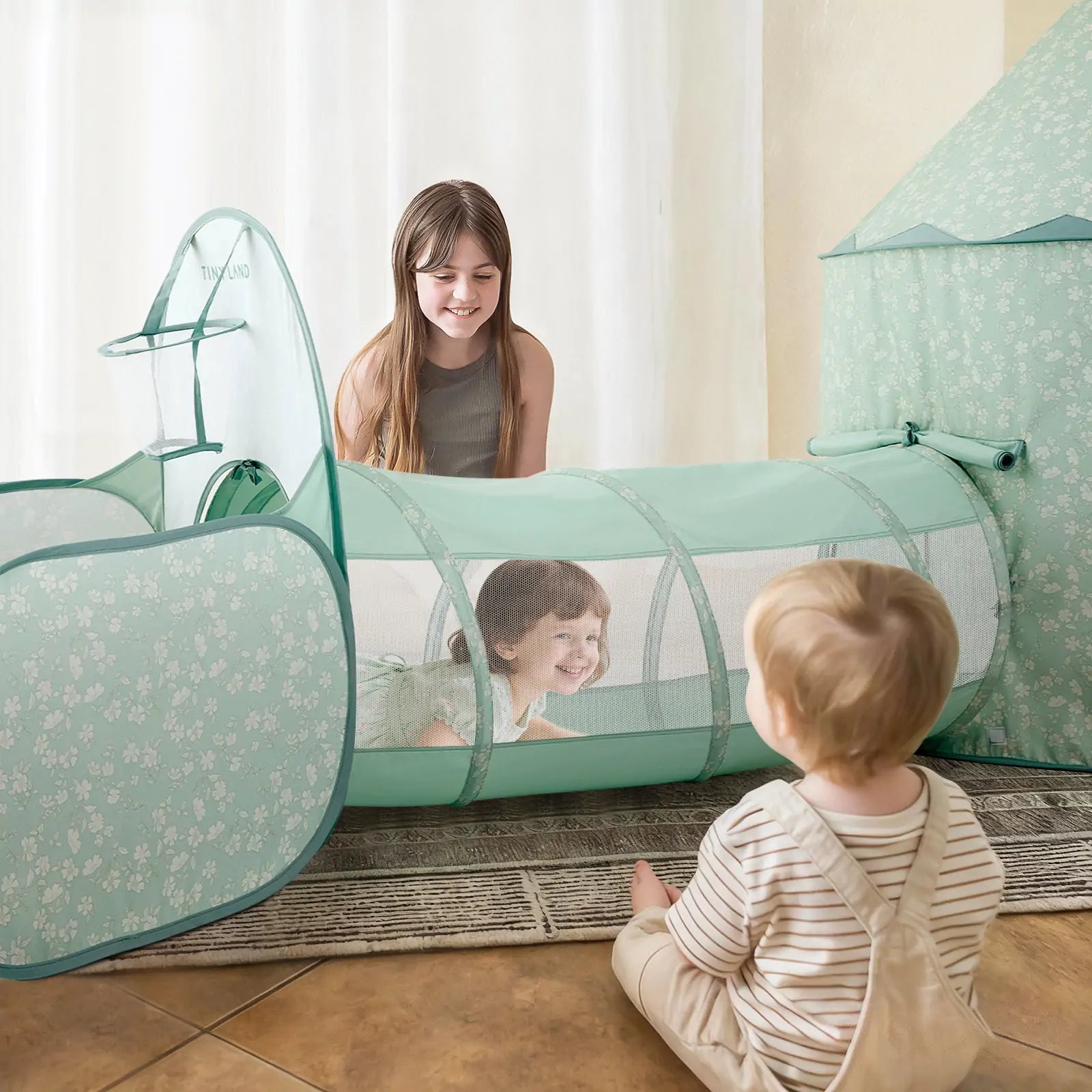 A young girl smiles at a toddler crawling through the mint green Tiny Land® Compact Adventure 3-in-1 Play Tunnel, while another toddler in striped overalls sits nearby. The Tiny Land set includes floral patterns, a matching playhouse, and a fun ball pit backdrop.