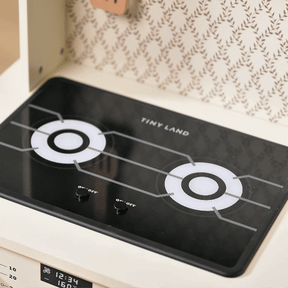 A close-up of the Tiny Land® PopJoy Play Kitchen shows two burners with circular designs and two black knobs, set against a patterned wall background. Brand: Tiny Land.