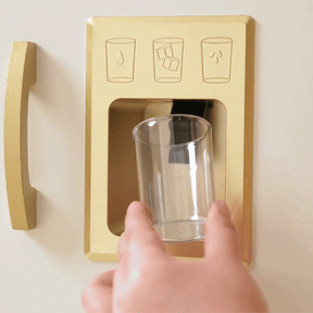 A hand holds a glass of colorful candies under the Tiny Land® PopJoy Play Kitchen fridge dispenser, where water and ice icons are shown above, blending playful imagination with realistic kitchen features.