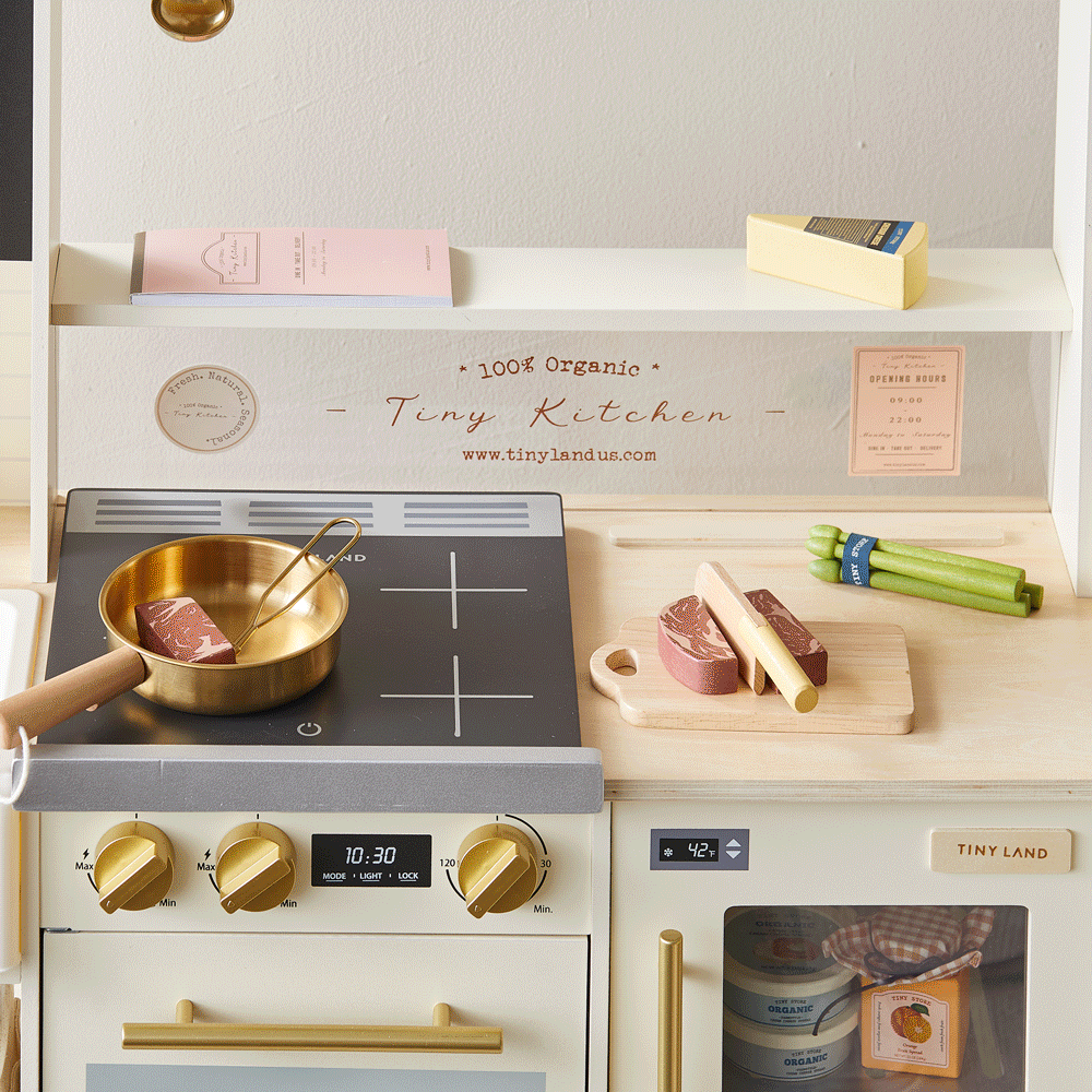 The Tiny Land® BistroSwitch Play Kitchen by Tiny Land features a stovetop, gold pan, cutting board, wooden food, pretend butter, an oven with fake food inside, shelves for accessories, and a pink notepad—perfect for endless pretend cooking fun.|White