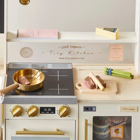 The Tiny Land® BistroSwitch Play Kitchen by Tiny Land features a stovetop, gold pan, cutting board, wooden food, pretend butter, an oven with fake food inside, shelves for accessories, and a pink notepad—perfect for endless pretend cooking fun.|White