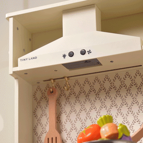 A close-up of the Tiny Land® PopJoy Kitchen & Fridge Set shows the range hood with buttons and decorative wallpaper. A wooden spoon and toy vegetables rest on the shelf, making this Tiny Land set ideal for imaginative play.