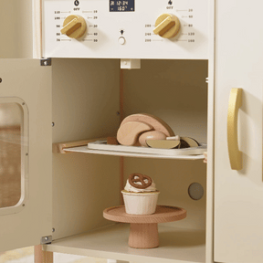The Tiny Land® PopJoy Play Kitchen oven, designed by Tiny Land, features cream and gold details and comes with wooden toy food: a tray of bread on the middle shelf and a cupcake with a pretzel topper on the bottom—ideal for any kids play kitchen.