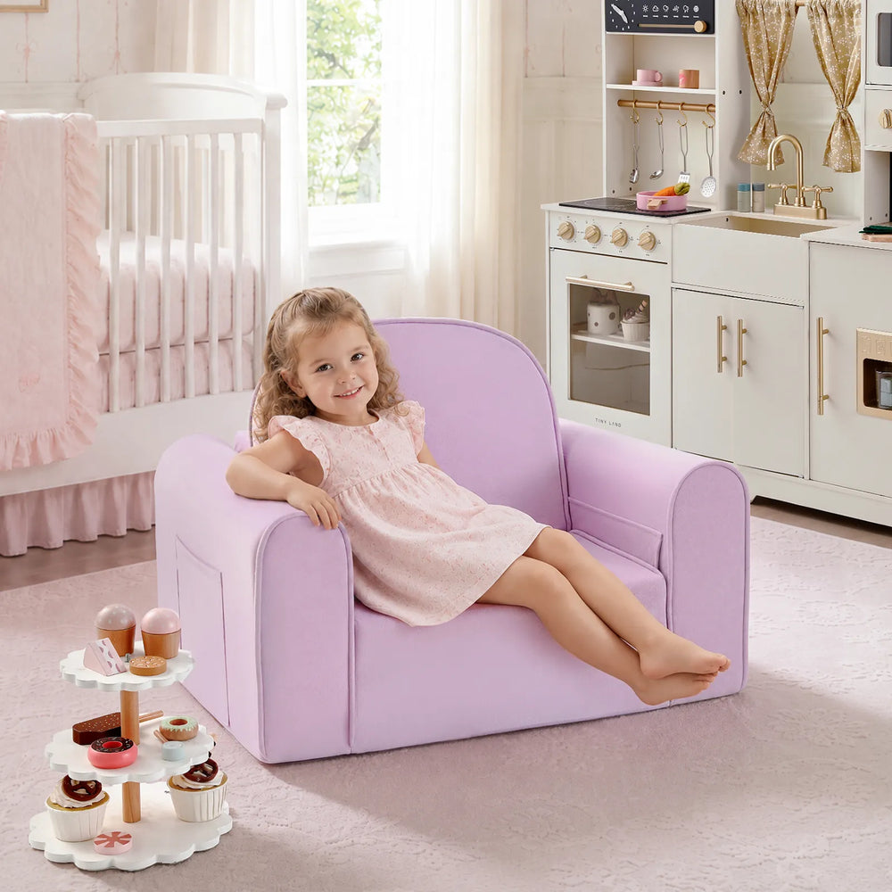 A young girl in a pink dress smiles while sitting on the Tiny Land® GentleSnug Kids Sofa Chair by Tiny Land—a light purple, flip-open seat—surrounded by adorable playroom furniture in a nursery with a crib, play kitchen, and toy dessert stand nearby.|Light Purple