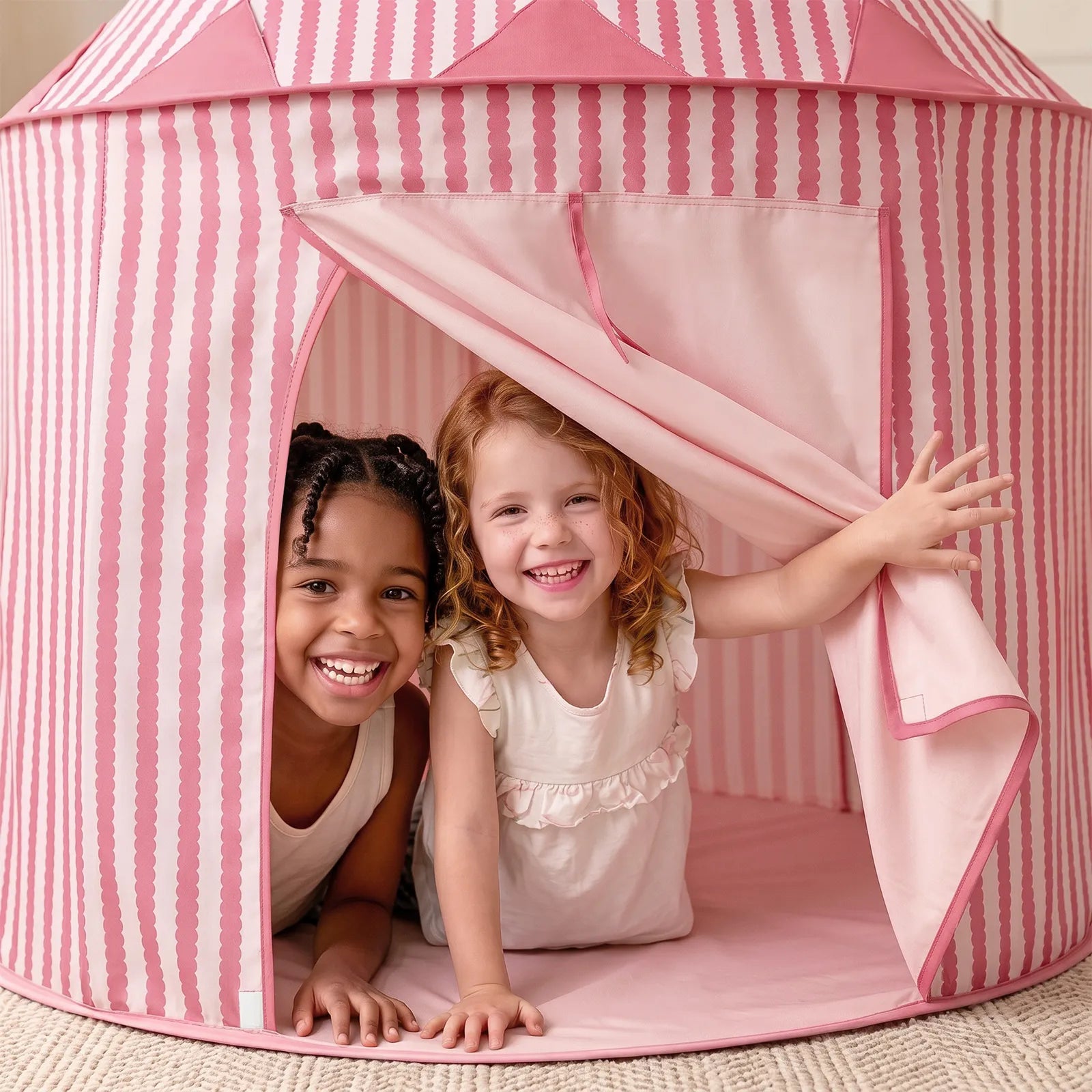 Two smiling young girls play inside the Tiny Land® Kids Tent – Indoor Tent for Toddlers by Tiny Land, with one holding the pink-and-white striped tent flap open. Both are wearing light-colored dresses and appear happy and playful.|Pink