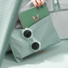 A hand with teal nails holds a tablet showing a rabbit, partially tucked into a mint-green striped pocket beside mint-green flower sunglasses and sand—perfect for playtime with the Tiny Land® 4-in-1 Baby Beach Tent & Sand Toy Set by Tiny Land.|Green