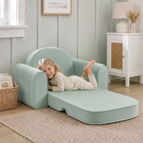A young child smiles while lying on the Tiny Land® GentleSnug Kids Sofa Chair in a cozy, softly lit room with neutral decor, a woven rug, small cabinet, and a basket filled with books nearby.
