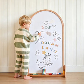 A young child in green and white striped pajamas draws on the Tiny Land® Double-Sided Arched Magnet Chalkboard, decorated with colorful magnetic letters, clouds, stars, and a smiling lion wearing a crown.